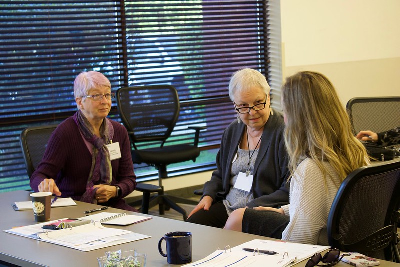 Three women have a discussion in a meeting room setting.